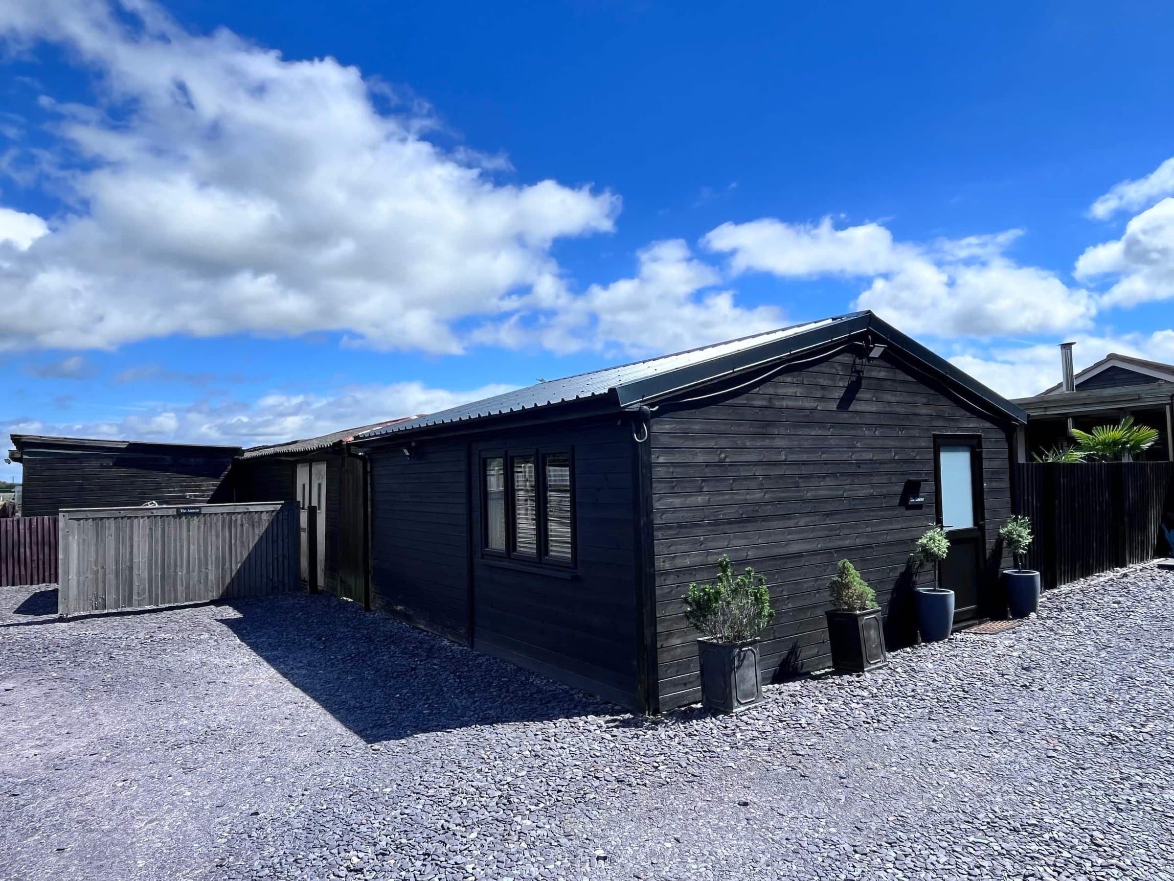The Annexe from the gravel parking area: a black timber building with pots of herbs by the door